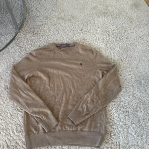 Beige stickad tröja Morris - Snygg beige stickad tröja från Morris med rund hals och diskret mörkblå logga broderad på bröstet. Tröjan har långa ärmar och ribbade muddar vid ärmslut och nederkant. Perfekt för dig som gillar stilrena och klassiska plagg.