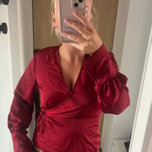 Vinröd omlottblus med knyt - Säljer en vinröd omlottblus med långa puffiga ärmar och knyt i sidan. Blusen har v-ringning och är tillverkad i ett glansigt, mjukt tyg som ger en lyxig känsla. Perfekt för dig som vill ha en trendig och feminin look.