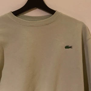 Beige sweatshirt från Lacoste - Snygg beige sweatshirt från Lacoste i storlek S. Klassisk rund hals och broderad grön krokodil på bröstet. Tröjan är gjord i mjuk bomull och har ribbade muddar vid halsen. Perfekt för chill dagar och enkel att matcha med jeans eller shorts.