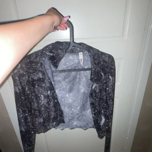 Svart mesh kofta med sol- och månprint - Säljer en svart mesh kofta från FB Sister i storlek XXS. Koftan har ett unikt vitt sol- och månprint över hela plagget, långärmad och med krage. Materialet är tunt och transparent, perfekt för att lägga till en cool touch till din outfit.