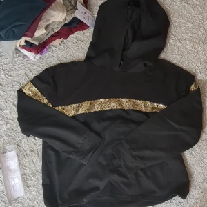 Svart hoodie med guldpaljetter - Svart hoodie med huva och lång ärm. Snygg detalj med ett brett band av glittrande guldpaljetter tvärs över bröstet och ut på ärmarna. Perfekt för dig som vill sticka ut lite extra och gillar en sportig men ändå glam stil. Har liksom genomskinligt material vid bytet☺️