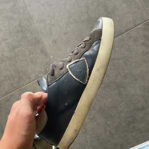 Philippe Model blå sneakers i skinn och mocka - Snygga sneakers från Philippe Model i mörkblått skinn med grå mockadetaljer och vit sula. Klassisk låg modell med snörning och rund tå. Ikonisk sköldlogga på sidan och bekväm platt sula. Perfekta för en stilren vardagslook. Dem är mustiga i bild men tvättar den såklart innan jag fraktar dem. Inte fast pris!!!!!!