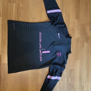 PSG x Nike svart half zip träningströja - Svart långärmad träningströja från Nike och PSG med rosa detaljer och broderad klubbmärke på bröstet. Tröjan har half zip, mönstrad struktur och rosa Nike-logga samt texten 'Accor Live Limitless' framtill. Perfekt för fotbollsträning eller chill. Byxornas högra dragkedja går inte att stänga men annars är den helt felfri.                                            Tröjan = 300kr   byxorna = 200kr  Set = 450