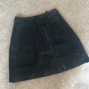 Svart skinnkjol med stora fickor - Snygg svart kort skinnkjol med två stora, markerade fickor framtill.