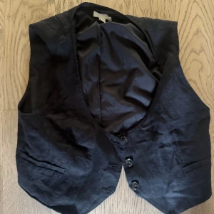 Svart kostymväst från H&M - Klassisk svart väst från H&M i storlek S. Västen har tre knappar framtill, två fickor och en snygg v-ringning. Perfekt att styla över en skjorta för en clean look. Materialet är slätt och västen har en figurnära passform.