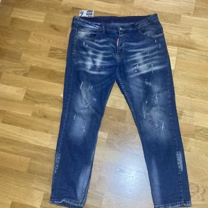 Dsquared2 blå jeans med slitningar - Snygga blå jeans från Dsquared2 med coola slitningar, färgstänk och en tvättad look. Klassisk femficksmodell med rakt snitt och röda detaljer vid gylfen. Jeansen har en modern vibe och passar perfekt till streetwear-stilen.