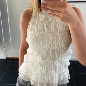 Vit omlott topp med knyt i ryggen - Supersöt vit omlott topp med tunna knytband i ryggen och halterneck. Toppen har ett plisserat tyg med volangkant nertill och är ärmlös. Perfekt för varma dagar när du vill ha en luftig och trendig look.