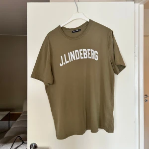 Olivgrön t-shirt från J.Lindeberg - Snygg olivgrön t-shirt från J.Lindeberg med vit logga tryckt över bröstet. Klassisk rund halsringning och korta ärmar. Tillverkad i mjuk bomull som känns skön mot huden. Perfekt för en clean och avslappnad stil. Köptes på Lindeberg för 699kr 