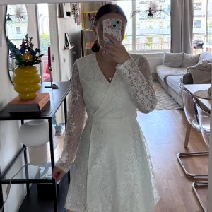 Vit spetsklänning från NLY Trend - Supersöt vit kortklänning från NLY Trend med omlottliv, v-ringning och långa ärmar i spets. Klänningen har markerad midja och är helfodrad förutom ärmarna som är transparenta. Perfekt för student eller sommarens alla tillfällen!