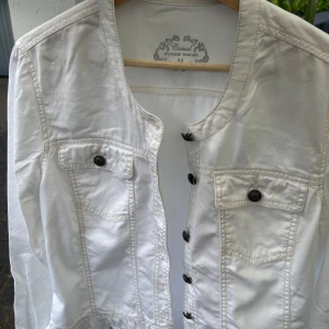 Vit jeansjacka från Mywear Woman - Snygg vit jeansjacka från Mywear Woman i klassisk modell med rund halsringning och svarta knappar framtill. Jackan har två bröstfickor med lock och knapp, samt markerade sömmar för en cool look. Perfekt att slänga över en t-shirt eller hoodie.