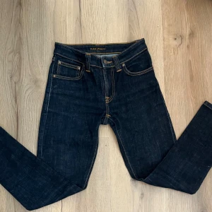 Mörkblå skinny jeans från Nudie Jeans - Snygga mörkblå jeans från Nudie Jeans med klassisk femficksdesign och kontrastsömmar. Modellen är skinny fit och har en smal passform hela vägen ner. Tillverkade i slitstarkt jeanstyg med orange detaljer och knappstängning framtill. Priset kan gå ner till 99kr om du följer oss på Plick och TikTok 