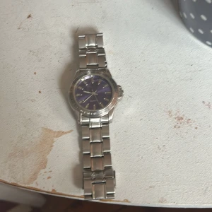 Silverfärgad klocka med lila urtavla - Stilren klocka från Quartz med metallarmband i silver och en snygg lila urtavla. Klockan har klassisk rund form, markerade index och detaljerad boett. Perfekt accessoar för dig som vill sticka ut med en cool färg på handleden.