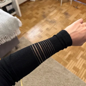 Svart tight stickad klönning med ränder - Säljer en svart, tight och stickad tröja med lång ärm. Klänningen har transparenta ränder över bröstet och på ärmarna som ger en snygg och modern look. Perfekt för dig som gillar stilrena plagg med en twist.