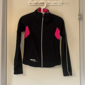 Svart och rosa vindjacka från Woxo - Sportig svart vindjacka från Woxo 720° med schyssta rosa detaljer på sidorna och insidan av kragen. Jackan har hel dragkedja framtill, långärmade ärmar med tumhål och reflekterande detaljer längs ärmarna. Perfekt för utomhusaktiviteter och träning.