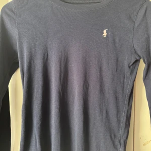 Mörkblå långärmad tröja Polo Ralph Lauren - Mörkblå långärmad tröja från Polo Ralph Lauren med klassisk rund hals och broderad logga i vitt på bröstet. Tröjan är i mjuk bomull och har en stilren, enkel design som passar perfekt till jeans eller kjol.