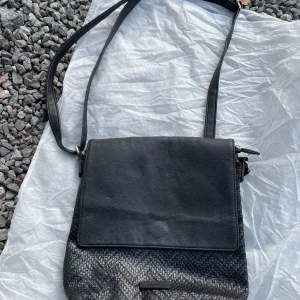 Svart axelväska från Don Donna - Snygg svart axelväska från Don Donna i skinnimitation med justerbar rem. Väskan har lock med strukturmönster och dragkedja upptill. Insidan har flera fack för att hålla ordning på dina grejer. Perfekt storlek för vardagens essentials.