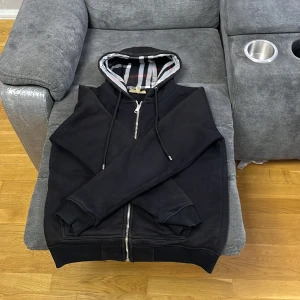 Burberry zip tröja  - Svart hoodiejacka från Burberry med dragkedja och klassiskt rutigt foder i huvan. Jackan har snörning i huvan och är tillverkad i mjuk bomull med lite elastan för extra komfort. Perfekt för dig som gillar stilren design med en touch av lyx. Storlek M 