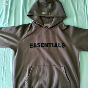 Mörkgrön Essentials hoodie med huva - Säljer en mörkgrön hoodie från Essentials Fear of God med svart tryck över bröstet. Tröjan har huva, känguruficka och en relaxed passform. Perfekt för dig som gillar streetwear och vill ha en stilren look.