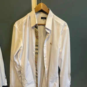 Vit Burberry skjorta XL med rutiga detaljer - Vit skjorta från Burberry i storlek XL men passar L med klassiska rutiga detaljer längs knappslån och på armbågarna. Skjortan har lång ärm, knappar framtill och är tillverkad i bomull. Perfekt för dig som gillar stilrena plagg med en twist.