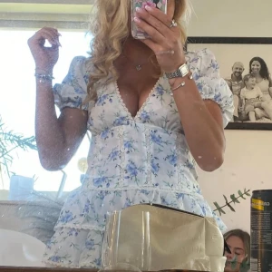 Blommig kortklänning gammal från Zara - Supersöt kortklänning i vitt med blått blommönster och puffärmar. Djup v-ringning och volangdetaljer ger en romantisk vibe. Perfekt för varma dagar och somriga tillfällen. Klänningen har en feminin passform och är tillverkad i ett lätt material.