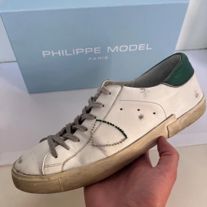 Philippe Model vita sneakers med gröna detaljer - Snygga vita sneakers från Philippe Model med gröna detaljer på hälen och sula. Skorna har grå snörning, rund tå och är tillverkade i skinn. Klassisk låg modell med diskret logga och kontrastfärgade sömmar på sidan.