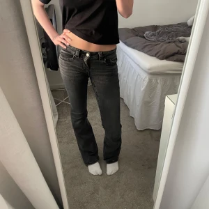 Svarta bootcut jeans med fickdetaljer - Snygga svarta jeans med bootcut passform och låg midja. Jeansen har coola fickdetaljer med knappar bak och klassisk femficksdesign. Perfekta för dig som gillar en lite retro vibe och vill ha ett par jeans som sticker ut.