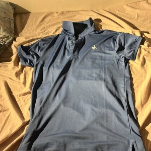 Blå pikétröja från Cross Sportswear - Snygg blå pikétröja från Cross Sportswear med klassisk krage och knappar framtill. Tröjan har korta ärmar och är tillverkad i ett mjukt och lätt polyestermaterial som andas. Perfekt för en sportig och avslappnad stil.