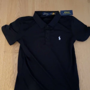 Svart pikétröja från Polo Ralph Lauren - Klassisk svart pikétröja från Polo Ralph Lauren med vit broderad logga på bröstet. Tröjan har korta ärmar, krage och knappar framtill. Tillverkad i mjuk bomull som känns skön mot huden. Perfekt för en stilren och sportig look.