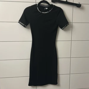 Svart kortklänning med textdetalj - Svart kortklänning med tight passform och korta ärmar. Runt halsringningen och ärmsluten finns en vit textdetalj med ordet 'ORIGINAL'. Enkel och stilren design som passar perfekt till sneakers eller boots.