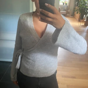 Ljusgrå omlott kofta med knyt - Supermysig ljusgrå stickad kofta med omlottmodell och knyt i sidan. Tröjan har lång ärm och är ribbad vid ärmslut och nederkant. Perfekt för lager-på-lager och chill dagar. Snygg och enkel att matcha med jeans eller kjol.