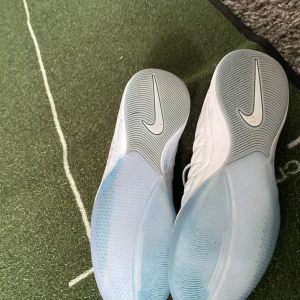 Nike Lunar Gato vita fotbollsskor - Nike Lunar Gato fotbollsskor i vitt med silverfärgad swoosh. Ovandel i mesh och skinn med quiltade detaljer framtill. Platt sula i genomskinlig ljusblå ton, snörning och sportig design. Perfekta för inomhusspel och futsal.