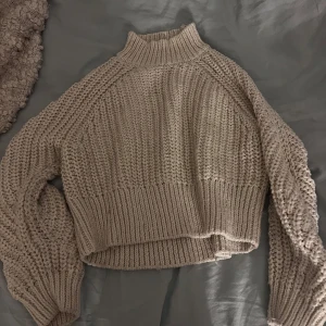 off-white stickad croppad tröja - Supermysig beige stickad tröja med croppad passform och hög krage. Tröjan har breda ribbade muddar vid ärmslut och nederkant samt grovstickat mönster. Perfekt för lager-på-lager och chill dagar.