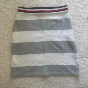 Randig kjol från Tommy Hilfiger - Snygg kort kjol från Tommy Hilfiger med breda grå och vita ränder. Midjan har en  resår med blå och röda detaljer. Perfekt för en fräsch och avslappnad look. Storlek 152 cm/12år
