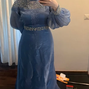 Blå långklänning med pärldetaljer - Elegant blå långklänning med transparenta puffärmar och vackra pärldetaljer vid halsringning, midja och ärmslut. Klänningen har en lätt glansig yta och markerad midja, perfekt för speciella tillfällen. Materialet känns lyxigt och ger en fin siluett. användt 2 gånger  passar M och L