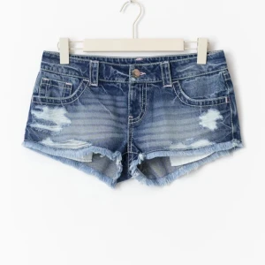 Lågmidjade jeansshorts med slitningar - Snygga blå jeansshorts från Gina Tricot med låg midja, slitna detaljer och fransig kant. Klassisk femficksmodell med coola nitar på bakfickorna. Perfekta för sommaren och ger en avslappnad vibe till din outfit.