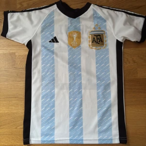 Argentina Messi 10 Adidas fotbollströja - Snygg Argentina fotbollströja från Adidas med klassiska ljusblå och vita ränder, svarta detaljer och broderade märken på bröstet. På ryggen står det Messi och nummer 10. Perfekt för dig som älskar fotboll och vill hylla en legend.