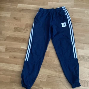 Blå Adidas mjukisbyxor med ränder - Snygga blå mjukisbyxor från Adidas med klassiska vita ränder längs sidorna och logga på låret. Byxorna har resår i midjan och vid bensluten, perfekt för en sportig och chill stil.