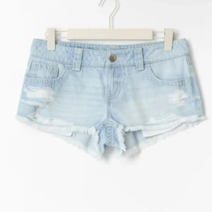 Ljusblå slitna jeansshorts Gina Tricot - Snygga ljusblå jeansshorts från Gina Tricot med låg midja, slitna detaljer och fransig kant. Shortsen har klassiska fickor fram och bak, samt coola nitar på bakfickorna. Perfekta för sommaren.