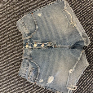 Blå jeansshorts med slitningar - Snygga blå jeansshorts med hög midja från Zara. Shortsen har slitna detaljer, 3 knappar framtill och klassiska fickor både fram och bak. Perfekta för sommaren och ger en avslappnad vibe.