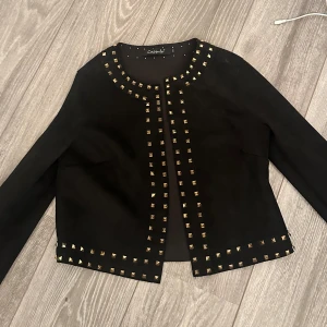 Svart kavaj med nitar från Capsule - Svart kavaj från Capsule med coola guldfärgade nitar längs krage, öppning och nederkant. Jackan har en rak, croppad passform och långa ärmar. Perfekt för dig som vill sticka ut med en edgy och trendig look. Jackan har svart mocka material 