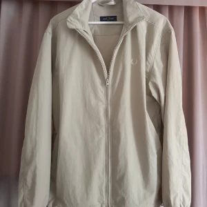 Beige vindjacka från Fred Perry - Snygg beige vindjacka från Fred Perry med klassisk krage och dragkedja framtill. Jackan har minimalistisk design med diskret broderad logga på bröstet och elastiska muddar. Perfekt för dig som gillar stilren och clean look. Använd 2-4 ggr, inga skador alls, precis som ny. Kvitto samt tags kommer med.