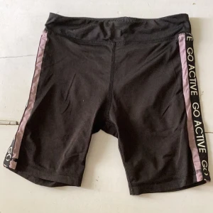 Svarta cykelshorts med lila detalj från KappAhl - Svarta cykelshorts från KappAhl med lila sidopanel och texten 'GO ACTIVE' längs benet. Stretchigt material som sitter tight och passar perfekt till träning eller sport. Bekväm midja och sportig look.