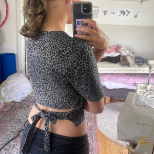 Leopardmönstrad croppad topp med knyt - Säljer en croppad topp med svart och grått leopardmönster. Toppen har korta ärmar och en snygg knytning i ryggen som ger en öppen detalj. Perfekt för dig som vill sticka ut med ett djurmönster och gillar trendiga detaljer.