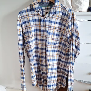 Rutig skjorta från Dressmann L - Snygg rutig skjorta från Dressmann i storlek L. Skjortan har blå, vita och gula rutor, klassisk krage och knäppning framtill. Tillverkad i mjuk bomull och har en bröstficka. Perfekt för en avslappnad och stilren look.