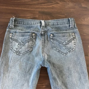 Ljusblå bootcut jeans med slitning - Säljer ett par ljusgrå jeans med bootcut passform och väldigt låg midja och klassiska femficksdetaljer. Jeansen har snygga slitningar och kontrastsömmar, samt bälteshällor och dragkedjegylf. Perfekta för dig som gillar en retro vibe och vill ha ett par jeans med lite extra attityd. Knaooen som man känner längts upp är borta men jag brukar sätta fast det med hårsnodd funkar toppen.