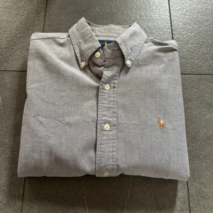 Ralph Lauren Skjorta - Grå (Stl S) - Säljer denna snygga gråa skjorta från Ralph Lauren. Skjortan är i gott skick men har en lagning längst ned på ryggen (bild 4) samt en mindre fläck (bild 5). Nypris ligger på ca 1400kr