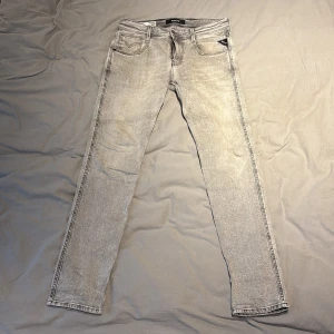 Replay Anbass grå slim jeans herr - Snygga grå jeans från Replay, modell Anbass. Slim fit med klassisk femficksdesign och svarta detaljer på bakfickorna. Jeansen har en ljusgrå tvätt och är tillverkade i mjukt denimtyg. Perfekta för dig som gillar en stilren och modern look.
