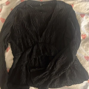 Svart topp från Gina Tricot - Säljer en svart topp från Gina Tricot i storlek L. Toppen har lång ärm, v-ringning och ett snyggt skrynklat tyg som ger en cool struktur. Perfekt för dig som gillar stilrena plagg med det lilla extra. Liten i storlek snarare som en M