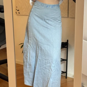 Ljusblå långkjol med broderi - Ljusblå långkjol i jeansliknande material med broderade blommor i midjan. Kjolen har dragkedja i sidan och en lätt utsvängd passform som ger en snygg siluett. Perfekt för dig som gillar en chill och stilren look. Saknar en knapp men enkelt att lösa