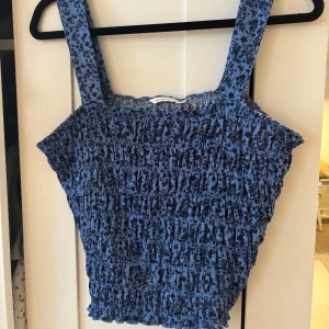 Blommig blå smockad linnetopp Dobber - Supersöt blå smockad linnetopp från Dobber med breda axelband och blommigt mönster. Toppen har en croppad passform och är perfekt för varma dagar. Materialet är mjukt och stretchigt för en bekväm känsla.